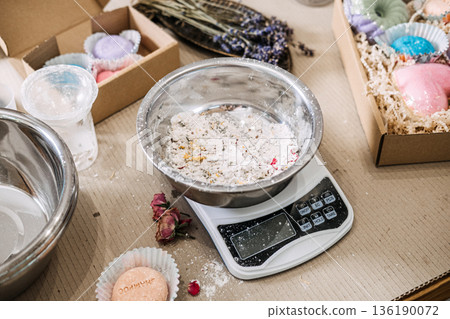 Metal bowl containing white powder and botanical inclusions sits on digital scale surrounded by packaging boxes. Cosmetic raw materials, ingredient study, clean formulation, skincare transparency. Metal bowl containing white powder and botanical inclusions sits on digital scale surrounded by packaging boxes. Cosmetic raw materials, ingredient study, clean formulation, skincare transparency. 136190072