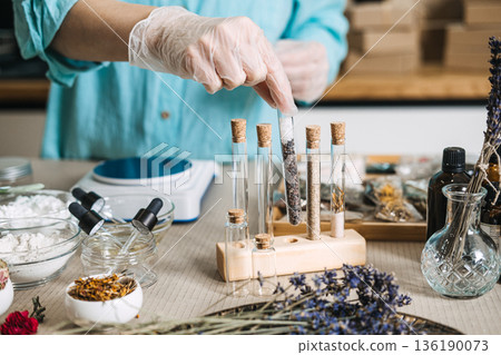 Gloved hand selects glass test tube filled with minerals and botanicals from wooden rack. Mood-regulating bath, cortisol rescue, stress-relief ritual, aromatherapy vial. 136190073