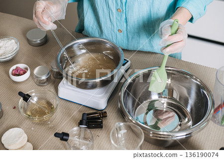Person uses glass rod to stir liquid in metal bowl on digital scale while holding green spatula. Precise cosmetic formulation process, aesthetic beauty lab scale, artisan skincare manufacturing. 136190074