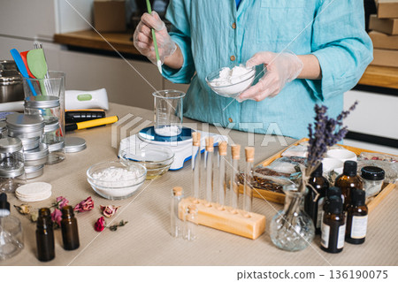 Person adds white powder from bowl into glass beaker on electronic scale surrounded by botanical ingredients. Lab-style skincare, botanical chemistry, diy cosmetic formulation, apothecary jars. 136190075