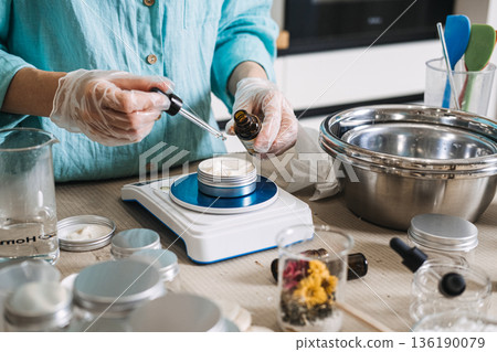 Person uses glass dropper to add oil to white cream in metal jar on digital scale. Lab-style beauty aesthetic, precise cosmetic formulation, diy apothecary lab, scientific skincare diy. 136190079