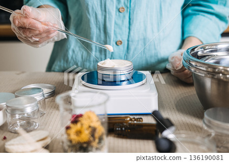 Person in gloves uses glass rod to apply cream into metal tin on digital scale. Botanical ingredients aesthetic, raw beauty materials, herbal mixology, natural skincare components. 136190081