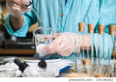 Person in gloves uses glass pipette to dispense liquid into beaker with white powder on digital scale. lab-style skincare, botanical chemistry aesthetic, diy cosmetic formulation, apothecary jars. 136190082