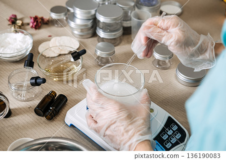 Person uses glass rod to stir white liquid in glass beaker on digital scale. Molecular skincare formulation, active ingredient dissolving, functional beauty craft, precision cosmetic measurement. 136190083