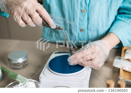 Gloved hands pour dark botanical salts from plastic packet into glass test tube on digital scale. Botanical layering, multi-salt DIY, herbal infusion shot, aesthetic bath salts. 136190085