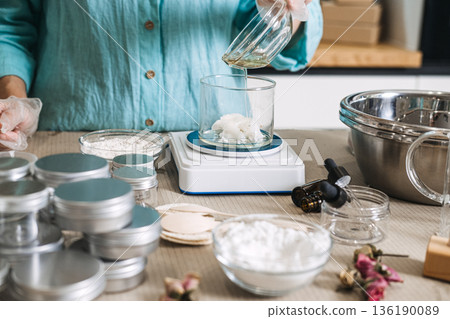 Hand in glove pours liquid oil from bowl into beaker containing white solid butter on digital scale. cosmetic formulation process, oil phase emulsion, cream texture transition, handmade moisturizer. 136190089
