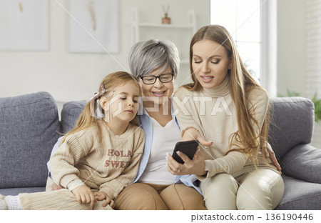 Happy grandmother, mother and granddaughter looking at smartphone screen and enjoying time together. 136190446