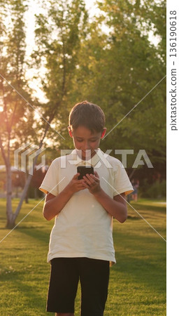Cheerful boy holds mobile phone in hands on green grass. Laughing boy chats with friends online. Cute boy watches funny video content. Social media communication. Internet surfing concept. Happy child Cheerful boy holds mobile phone in hands on green grass. Laughing boy chats with friends online. Cute boy watches funny video content. Social media communication. Internet surfing concept. Happy child 136190618