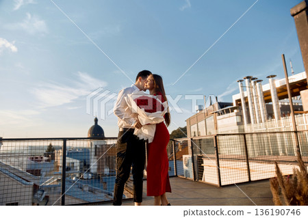 Couple shares a kiss on a rooftop at sunset while holding a bouquet of red roses in the city Couple shares a kiss on a rooftop at sunset while holding a bouquet of red roses in the city 136190746