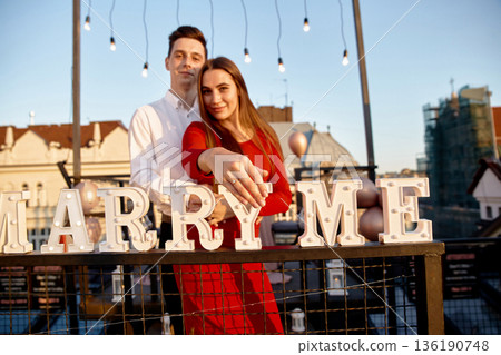 Couple enjoys engagement moment on rooftop during sunset with Marry Me letters in background 136190748