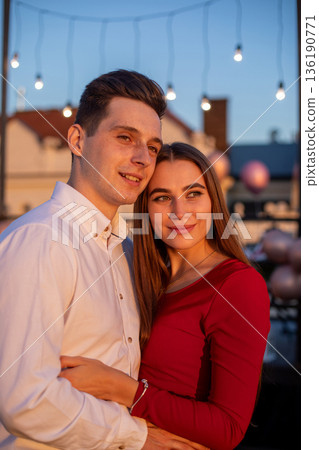 Couple stands close together on a rooftop during sunset while string lights hang above in the background 136190771