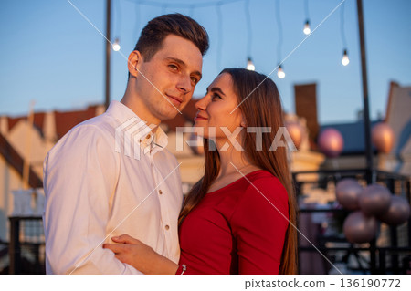Couple stands close on a rooftop during sunset with lights hanging around them and balloons in the background Couple stands close on a rooftop during sunset with lights hanging around them and balloons in the background 136190772