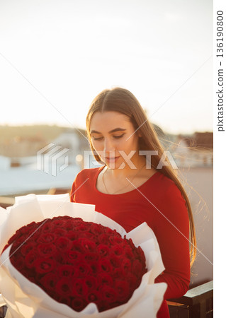 Woman holds large bouquet of red roses in outdoor setting during sunset 136190800