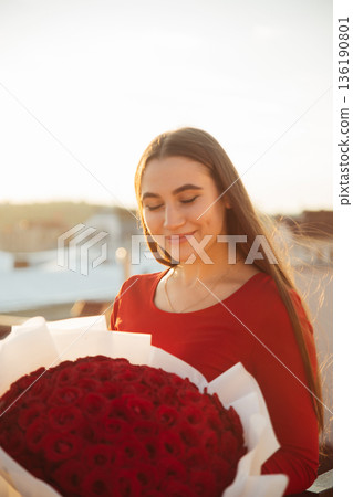 Woman holds large bouquet of red roses while standing on rooftop during sunset in the city Woman holds large bouquet of red roses while standing on rooftop during sunset in the city 136190801