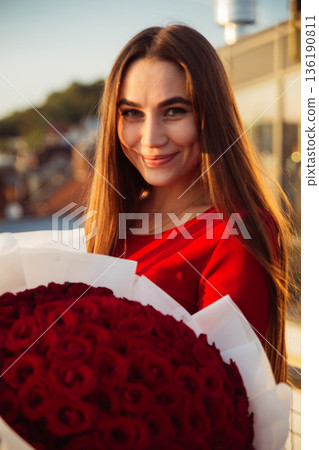 Woman holding a large bouquet of red roses on a sunny day while smiling in an outdoor setting 136190811