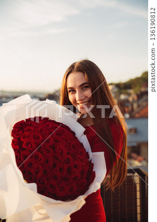 Woman smiles while holding large bouquet of red roses on balcony during sunset in city Woman smiles while holding large bouquet of red roses on balcony during sunset in city 136190812