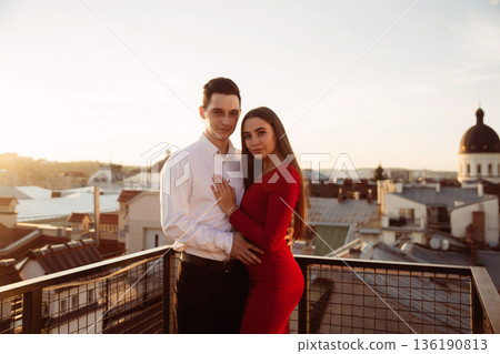 Couple stands together on a rooftop during sunset with city buildings in the background Couple stands together on a rooftop during sunset with city buildings in the background 136190813