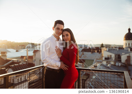 Couple stands together on rooftop during sunset overlooking city skyline Couple stands together on rooftop during sunset overlooking city skyline 136190814