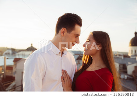 Two people share a moment on a rooftop at sunset near a city with buildings and water in the background 136190818