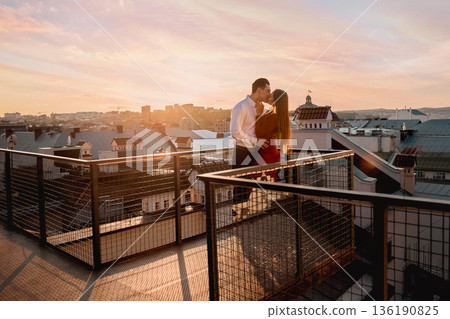 Couple shares a kiss on a rooftop during sunset in a city with varied architecture and buildings as the backdrop 136190825