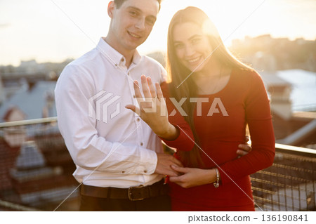 Couple shares moment on rooftop during sunset as they celebrate engagement and display ring to the camera with joy 136190841