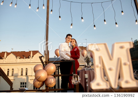 Couple celebrates special occasion on rooftop at sunset with lights and decorations in the background Couple celebrates special occasion on rooftop at sunset with lights and decorations in the background 136190992