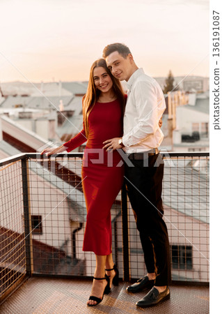 Couple poses on balcony at sunset in city, enjoying warm evening and each other company Couple poses on balcony at sunset in city, enjoying warm evening and each other company 136191087