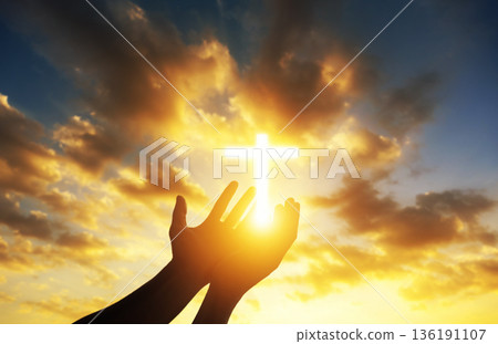 Human hands open palm up worshiping a glowing Christian cross in sunset sky. Concept for faith, spirituality, and hope. Human hands open palm up worshiping a glowing Christian cross in sunset sky. Concept for faith, spirituality, and hope. 136191107