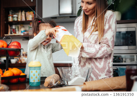 Mother and daughter work together in the kitchen making a recipe with flour and sugar while enjoying their time on a sunny afternoon 136191350