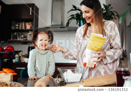 Woman and child prepare food in kitchen with flour and other ingredients while smiling and enjoying their time together during the day Woman and child prepare food in kitchen with flour and other ingredients while smiling and enjoying their time together during the day 136191352