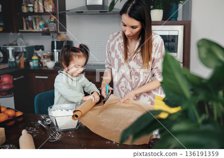 Mother and daughter work together on baking project in home kitchen during the afternoon preparing dough and cutting parchment paper Mother and daughter work together on baking project in home kitchen during the afternoon preparing dough and cutting parchment paper 136191359
