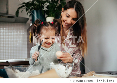 Mother and daughter cooking together in the kitchen while preparing food with eggs and ingredients on the table during a sunny morning 136191364