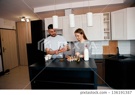 In a stylish kitchen with light colors, a couple joyfully cooks together. They mix ingredients in a bowl while sharing a tender moment, creating a warm atmosphere 136191387