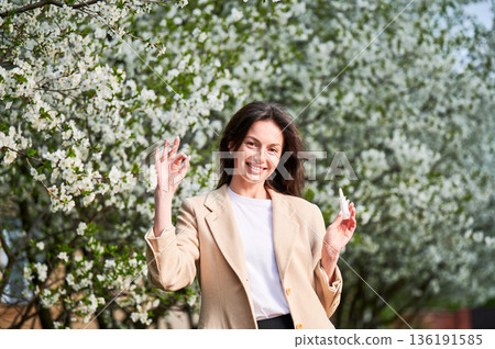 Woman allergic using medical nasal drops, suffering from seasonal allergy in blossoming garden. Smiling woman holding nasal spray, making 'OK' hand sign near blooming tree. Spring allergy concept. Woman allergic using medical nasal drops, suffering from seasonal allergy in blossoming garden. Smiling woman holding nasal spray, making 'OK' hand sign near blooming tree. Spring allergy concept. 136191585