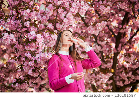 Woman allergic using medical nasal spray, suffering from seasonal allergy at spring in blossoming garden. Young woman treating runny nose in front of blooming tree outdoors. Spring allergy concept. Woman allergic using medical nasal spray, suffering from seasonal allergy at spring in blossoming garden. Young woman treating runny nose in front of blooming tree outdoors. Spring allergy concept. 136191587