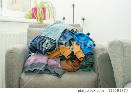 Stack of folded clothes placed on a gray armchair in a bright living room, everyday home routine, laundry concept, domestic life and organization. Stack of folded clothes placed on a gray armchair in a bright living room, everyday home routine, laundry concept, domestic life and organization. 136191874