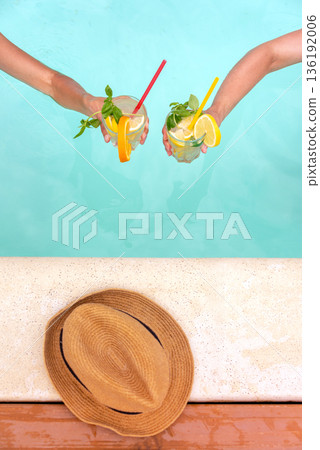 Two hands hold colorful cocktails garnished with fruits and mint over a blue pool. A straw hat rests on the pool edge. Perfect for depicting leisure, summer relaxation, and tropical vacation vibes. Two hands hold colorful cocktails garnished with fruits and mint over a blue pool. A straw hat rests on the pool edge. Perfect for depicting leisure, summer relaxation, and tropical vacation vibes. 136192006