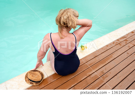 A woman in a swimsuit relaxes poolside. The serene setting with a refreshing beverage highlights summer leisure and tranquility, perfect for vacation and self-care concepts. A woman in a swimsuit relaxes poolside. The serene setting with a refreshing beverage highlights summer leisure and tranquility, perfect for vacation and self-care concepts. 136192007