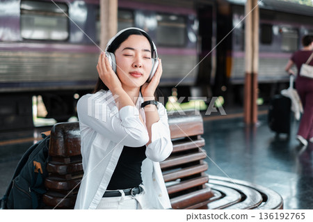 A young woman sitting on a bench at a train station, wearing headphones and enjoying music with eyes closed. 136192765