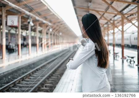 Woman with headphones waits at a train station, symbolizing modern travel and commuting. 136192766