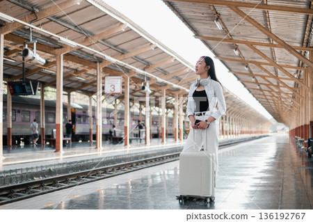 Woman standing with luggage on a train station platform, looking into the distance under a sunlit roof. 136192767