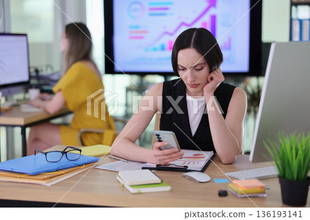 Busy office scene with focused woman checking mobile phone among papers and reports 136193141