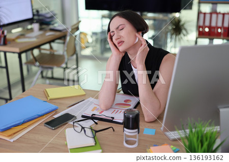 Woman experiencing stress during work hours in a modern office setting with paperwork scattered around 136193162