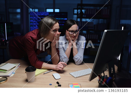 Two women in an office express concern while examining a computer screen during a late night work session Two women in an office express concern while examining a computer screen during a late night work session 136193185