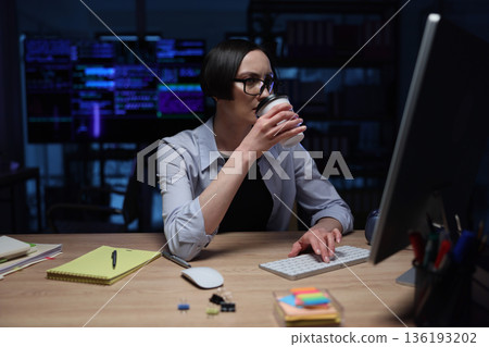 Focused programmer enjoys a warm drink while coding in a dimly lit office at night 136193202