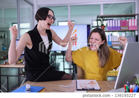 Two joyful women celebrating success in a modern office, exuding energy and teamwork, surrounded by colorful office supplies Two joyful women celebrating success in a modern office, exuding energy and teamwork, surrounded by colorful office supplies 136193227