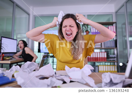 Woman expresses frustration in an office filled with crumpled papers and stressed coworkers during a busy workday 136193248