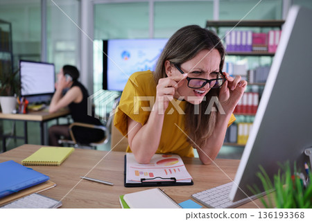 Busy office scene captures a woman focused on her work while her colleague is on the phone, surrounded by colorful charts and documents Busy office scene captures a woman focused on her work while her colleague is on the phone, surrounded by colorful charts and documents 136193368