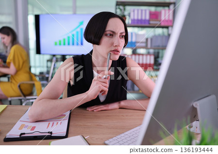 Focused business woman analyzing data in a modern office while another colleague works on a project in the background 136193444