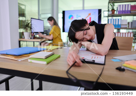 Busy office scene shows hardworking woman feeling exhausted while her colleague focuses on computer tasks 136193449